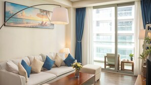 A living room with a sofa, coffee table, and a painting of a beach scene. - A la Carte - Coralie Luxury Seaview Aprtment (Đà Nẵng)