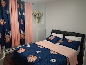 Une chambre à coucher avec un lit, des rideaux à motifs floraux, un vase avec des fleurs et un radiateur.