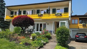 Un immeuble de deux étages avec un balcon jaune, une voiture garée devant et un jardin où poussent différentes plantes.