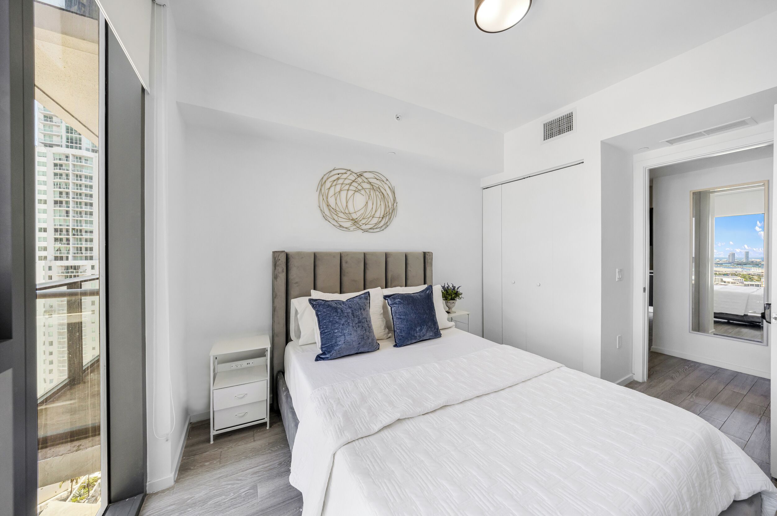 A bedroom with a bed, a nightstand, a wall art piece, and a view of the city through a window.