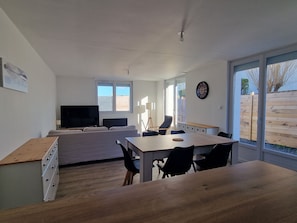 A modern living room with a sofa, TV, dining table, chairs, and a large window. - "Chez G & G" Renovated family home sleeps 8, walking distance to beach. (La Hague)