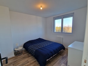 A bedroom with a bed, a small table, a radiator, and a window. - "Chez G & G" Renovated family home sleeps 8, walking distance to beach. (La Hague)