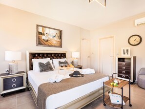Une chambre d’hôtel avec un lit, des tables de chevet, une petite table, une lampe, une horloge, un cadre photo, un plateau avec des verres et un chapeau, et un magazine sur une table d’appoint.