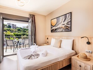 Ein Hotelzimmer mit Bett, Nachttisch, Wandlampe, einem Gemälde und Blick auf einen Balkon mit Gartenmöbeln.