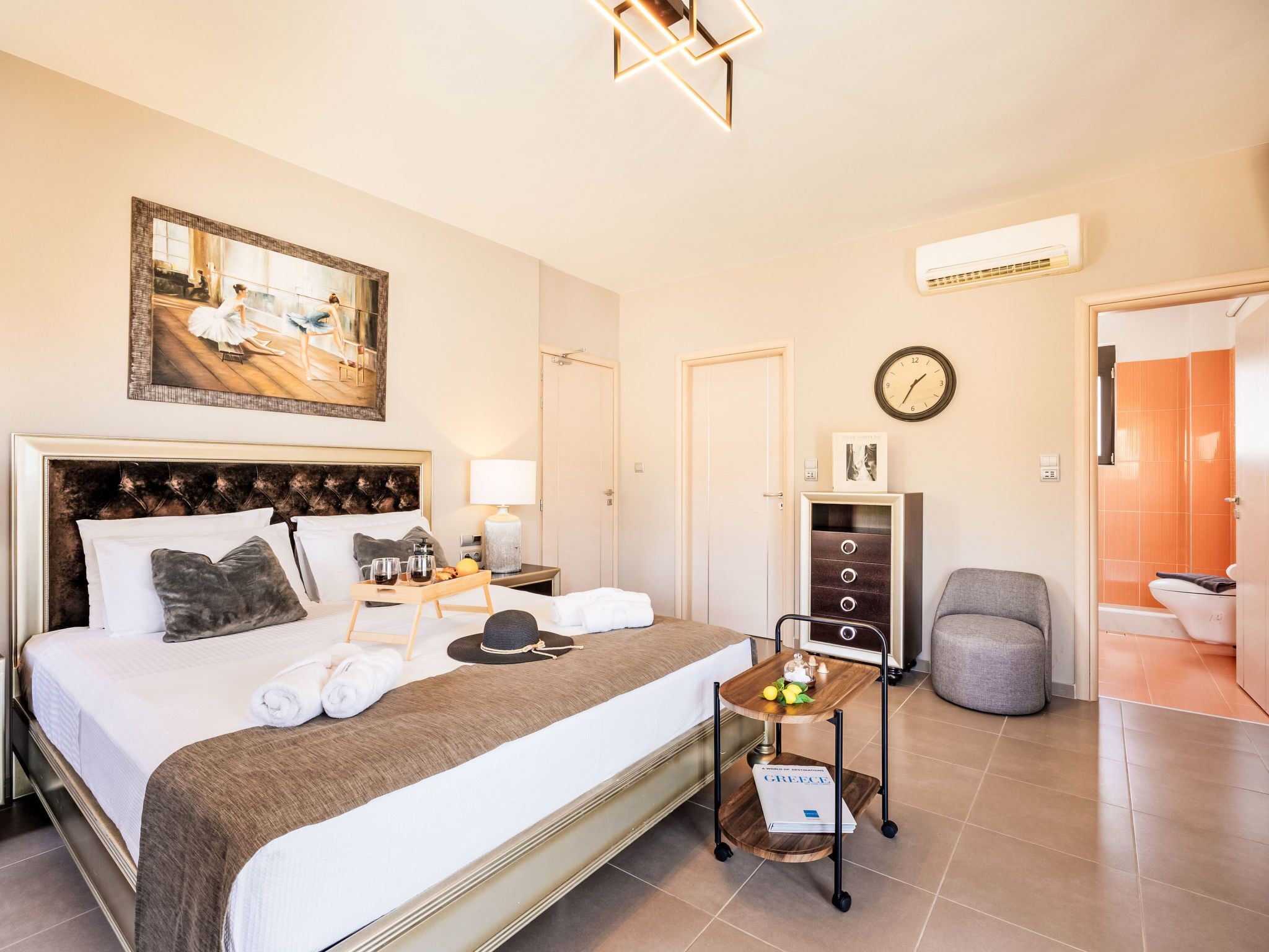 Quarto de hotel com uma cama grande, mesa de cabeceira com abajur, mesinha lateral com bandeja, relógio e porta para o banheiro.
