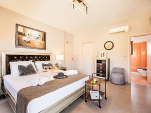 Quarto de hotel com uma cama grande, mesa de cabeceira com abajur, mesinha lateral com bandeja, relógio e porta para o banheiro.