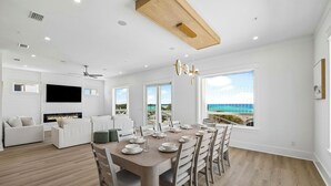 Ett modernt vardagsrum med en öppen spis, vita soffor, ett matbord för sex personer och stora fönster med utsikt över havet.