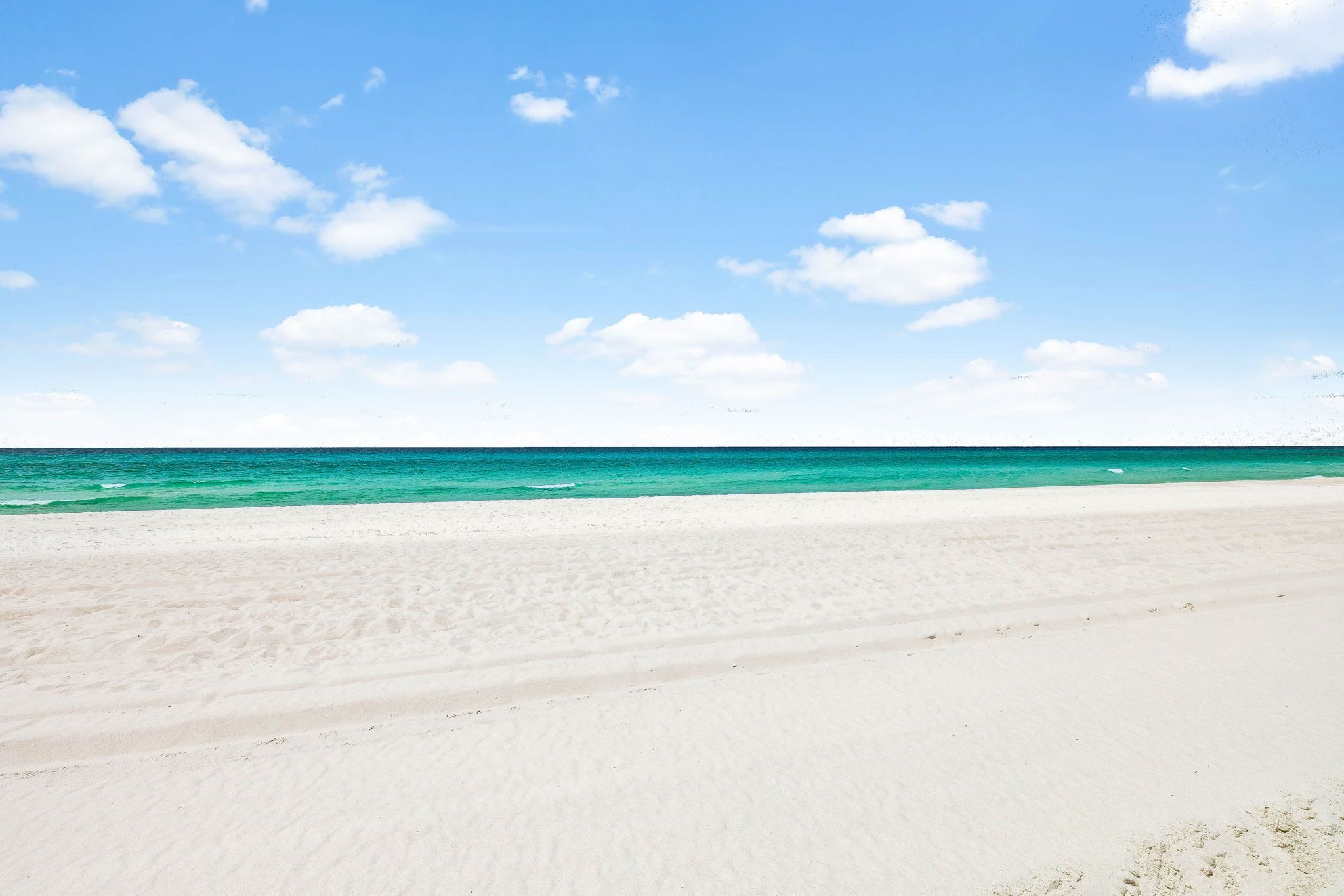 A beach with white sand and clear turquoise water under a blue sky with scattered clouds.