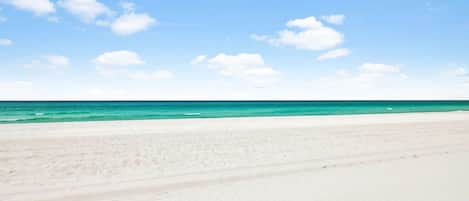 Een strand met wit zand en helder turquoise water onder een blauwe lucht met losse wolken.