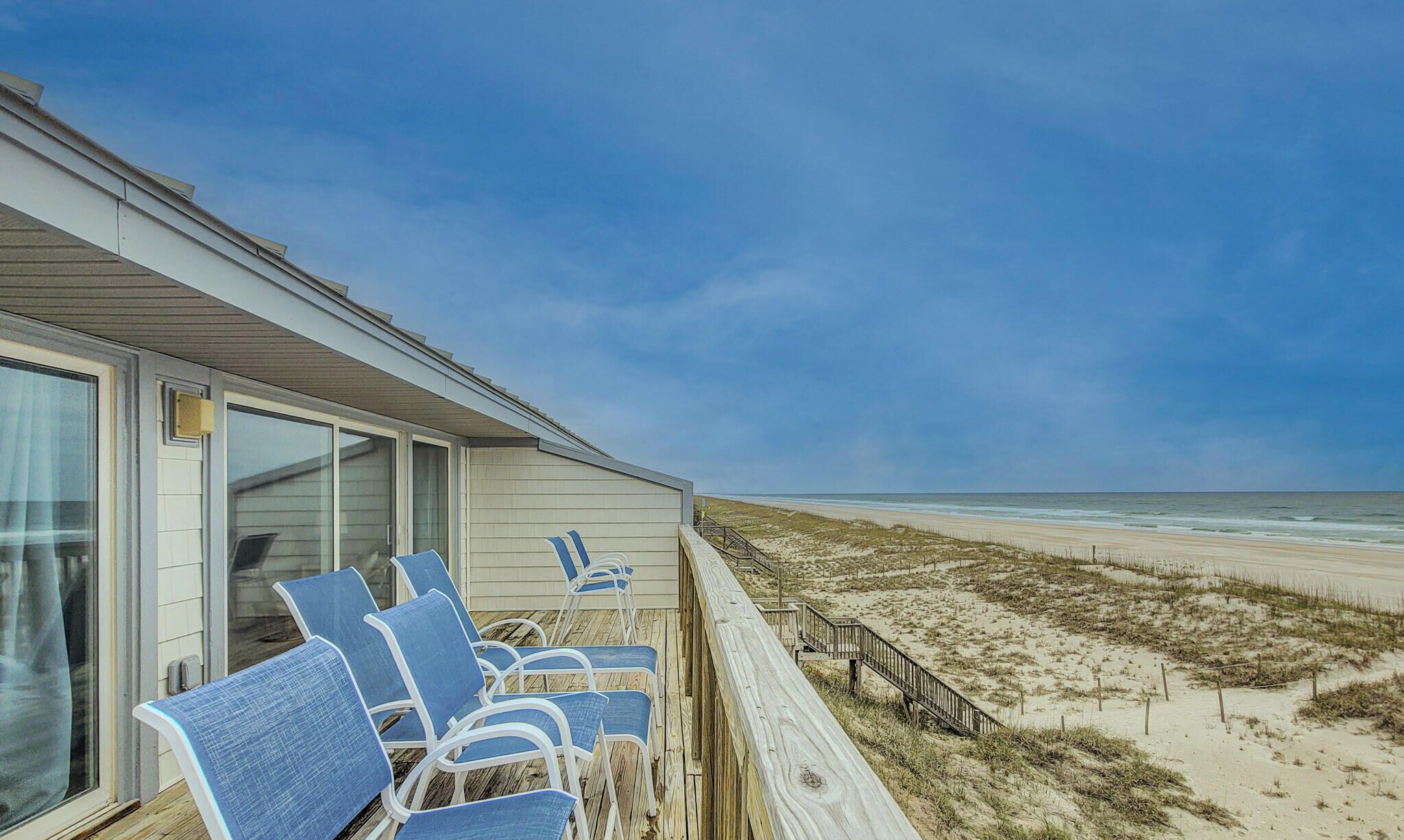 Een gebouw aan het strand met een terras, blauwe stoelen en een houten looppad naar het strand.