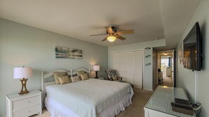Ein Schlafzimmer mit Bett, Nachttischlampen, Deckenventilator, Nachttisch, Sessel, Kommode, Spiegel und einer Tür zu einem anderen Raum.