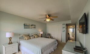 A bedroom with a bed, bedside lamps, a ceiling fan, a nightstand, a chair, a dresser, a mirror, and a door leading to another room. - Queen's Grant E-216 - NEW - Open Summer Weeks! (Topsail Beach)