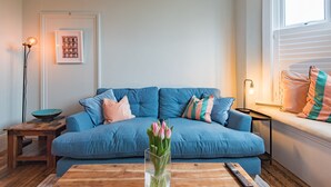 A living room with a blue sofa, a wooden coffee table, a vase of flowers, a framed picture on the wall, and a floor lamp. - Whispering Waves, Hythe (Hythe)