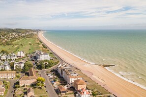 Vista aérea de un pueblo costero con una extensa playa de arena, edificios residenciales y una carretera que discurre paralela a la costa.