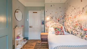A bedroom with a bed, a nightstand, a dresser, a mirror, a fireplace, and a floral wallpaper. - Whispering Waves, Hythe (Hythe)