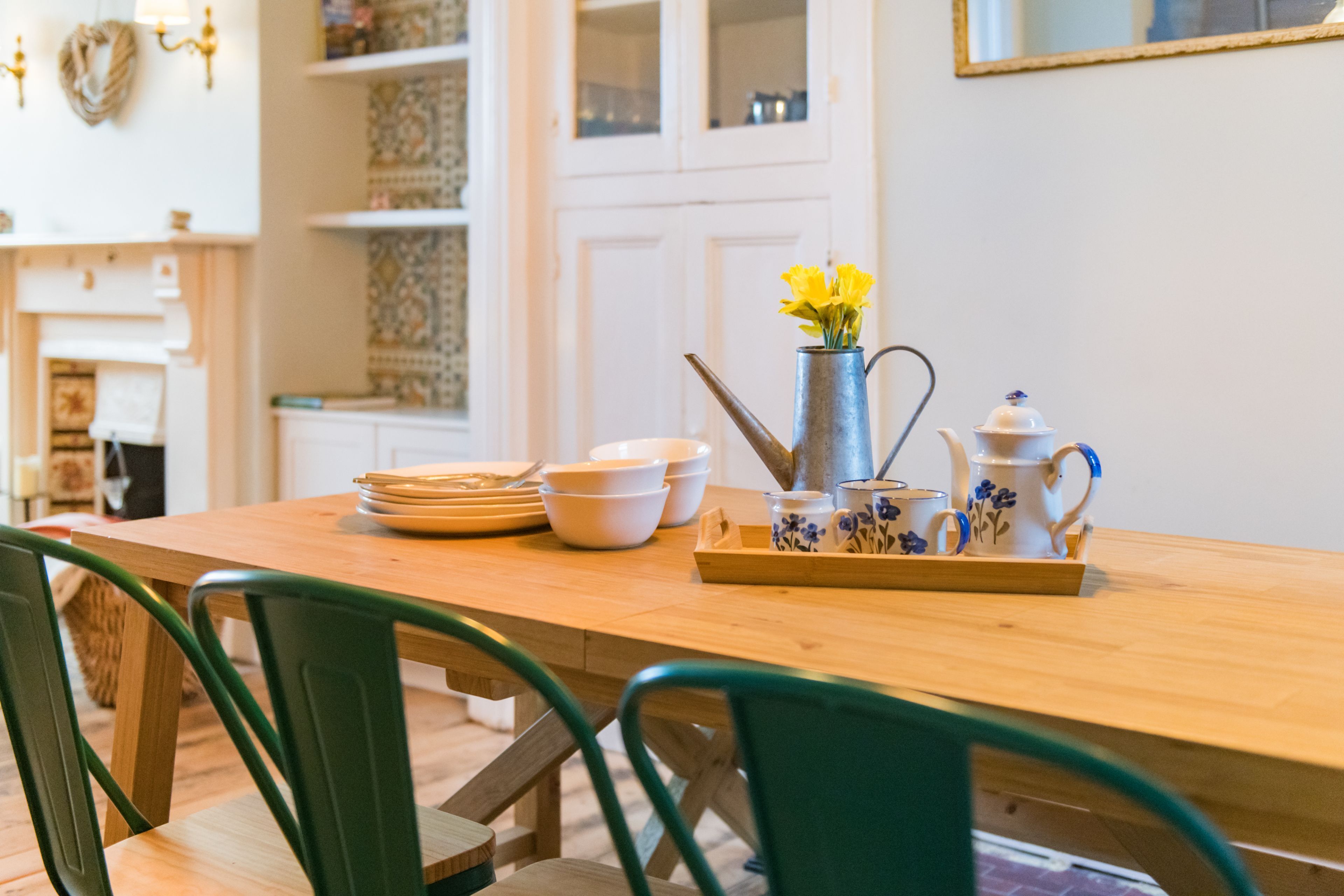 A wooden dining table with chairs, a vase with yellow flowers, and a tray with teacups and a pitcher.