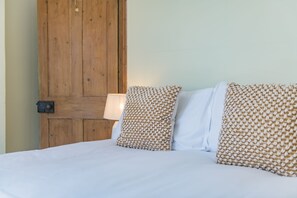 A bed with white bedding and patterned pillows, a wooden door, and a lamp on a bedside table. - Pebble Beach Cottage (Kingsdown)