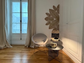 Une pièce avec un plancher en bois, une chaise blanche, une table ronde avec une lampe et un élément décoratif fixé au mur.