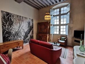 Un salon avec un canapé rouge, un meuble en bois, un tableau et une grande fenêtre avec des volets.