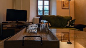 Uma sala de estar com televisão, uma mesa de vidro com copos de vinho, um banco de madeira e uma janela com vista.