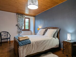 A bedroom with a wooden ceiling, a bed with white bedding, a chair, a nightstand with a lamp, and a window with curtains.