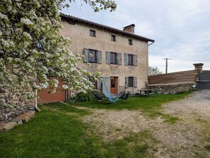 Un bâtiment à deux étages doté d’une façade en pierre, de nombreuses fenêtres avec volets, d’une porte en bois et d’un chemin de gravier menant à un espace aménagé avec des chaises en métal.