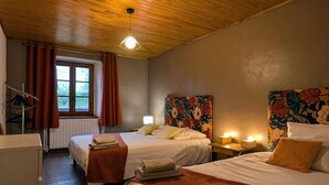 Quarto de hotel com duas camas, teto de madeira, janela com cortinas, radiador e mesa de cabeceira com abajur.