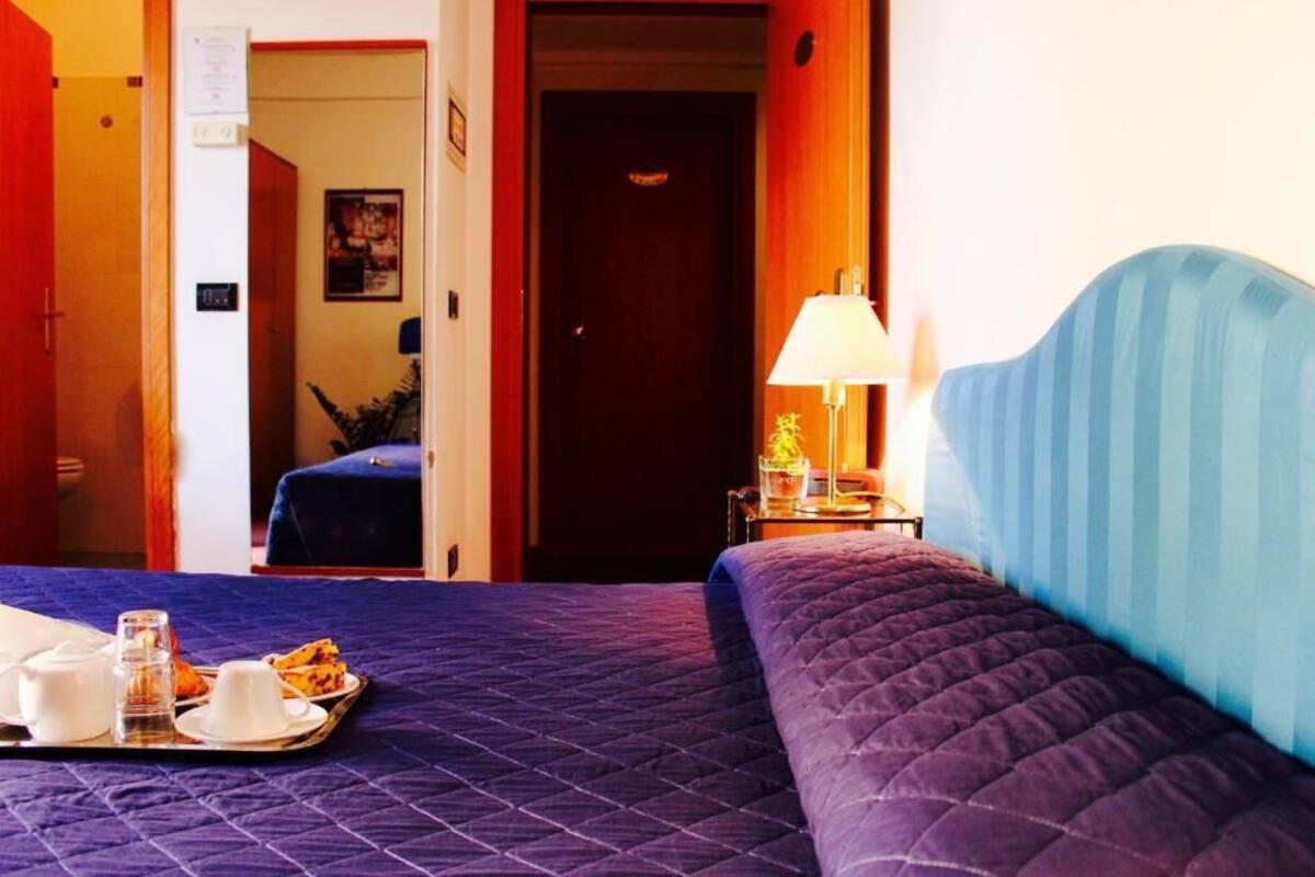 Una habitación de hotel con una cama, una bandeja con artículos de desayuno, una mesita de noche con una lámpara y una puerta que da a otra habitación.