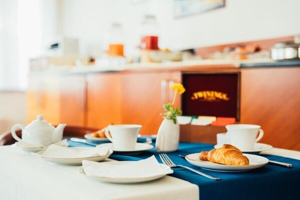 Una preparación para el desayuno con un croissant, tetera, tazas y un florero con una flor amarilla sobre una mesa.