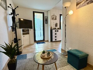 Ein kompakter Wohnbereich mit Flachbildfernseher, einer kleinen Essecke, einer Topfpflanze, einem runden Tisch mit dekorativer Tischdekoration und einer Stehlampe.