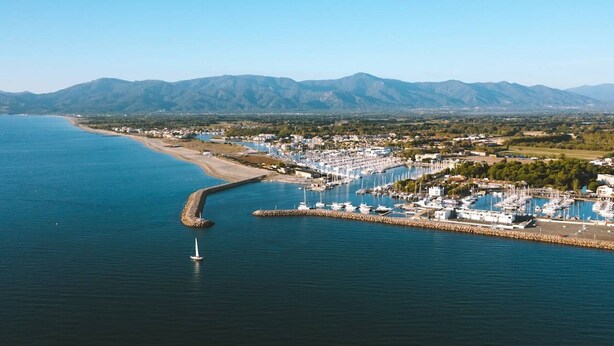 Una marina costanera amb vaixells, un moll i una població amb muntanyes al fons.