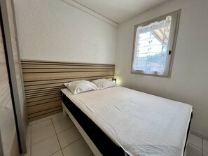 Ein Einzelbett mit Holz Kopfteil, weißer Bettwäsche und einem Fenster mit Blick auf ein Gebäude und Bäume.