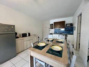 A compact kitchen with a refrigerator, microwave, and stove, a dining table set for two with plates and glasses, and a small cabinet with kitchen utensils. - Apartment with AC and parking - 3 guests (Saint-Cyprien)