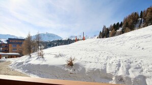 Pendio innevato con una ringhiera in legno in primo piano, un edificio con finestre e un tetto imbiancato di neve sullo sfondo, e un cielo sereno.