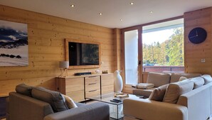 Een moderne woonkamer met houten details, een grote flatscreen televisie, een bank, een salontafel en uitzicht op herfstbomen door een groot raam.