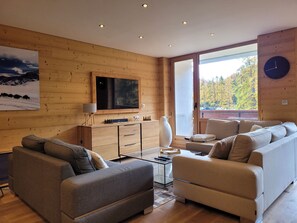 Una sala de estar moderna con detalles de madera, un televisor de pantalla plana grande, un sofá, una mesa de centro y vistas a árboles otoñales a través de un ventanal amplio.