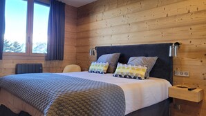 Een slaapkamer met een houten wand, een bed met een grijs hoofdeinde, twee bedlampjes, een nachtkastje met een lade en een raam met gordijnen.