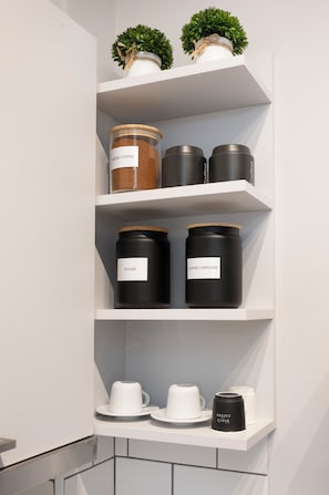 Ein Küchenregal mit Gläsern, die mit 'Griechischer Kaffee', 'Zucker' und 'Kaffeekapseln' beschriftet sind, sowie eine Kaffeemaschine auf einer weißen Arbeitsplatte.