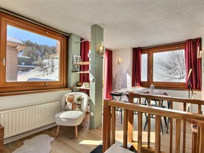 Ein gemütliches Zimmer mit Blick auf eine verschneite Landschaft, ein Holzsessel, ein Esstisch und eine Treppe.