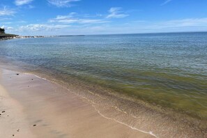 Ein Strand mit klarem Wasser, sandigem Ufer und einer entfernten Küstenlinie mit Gebäuden.