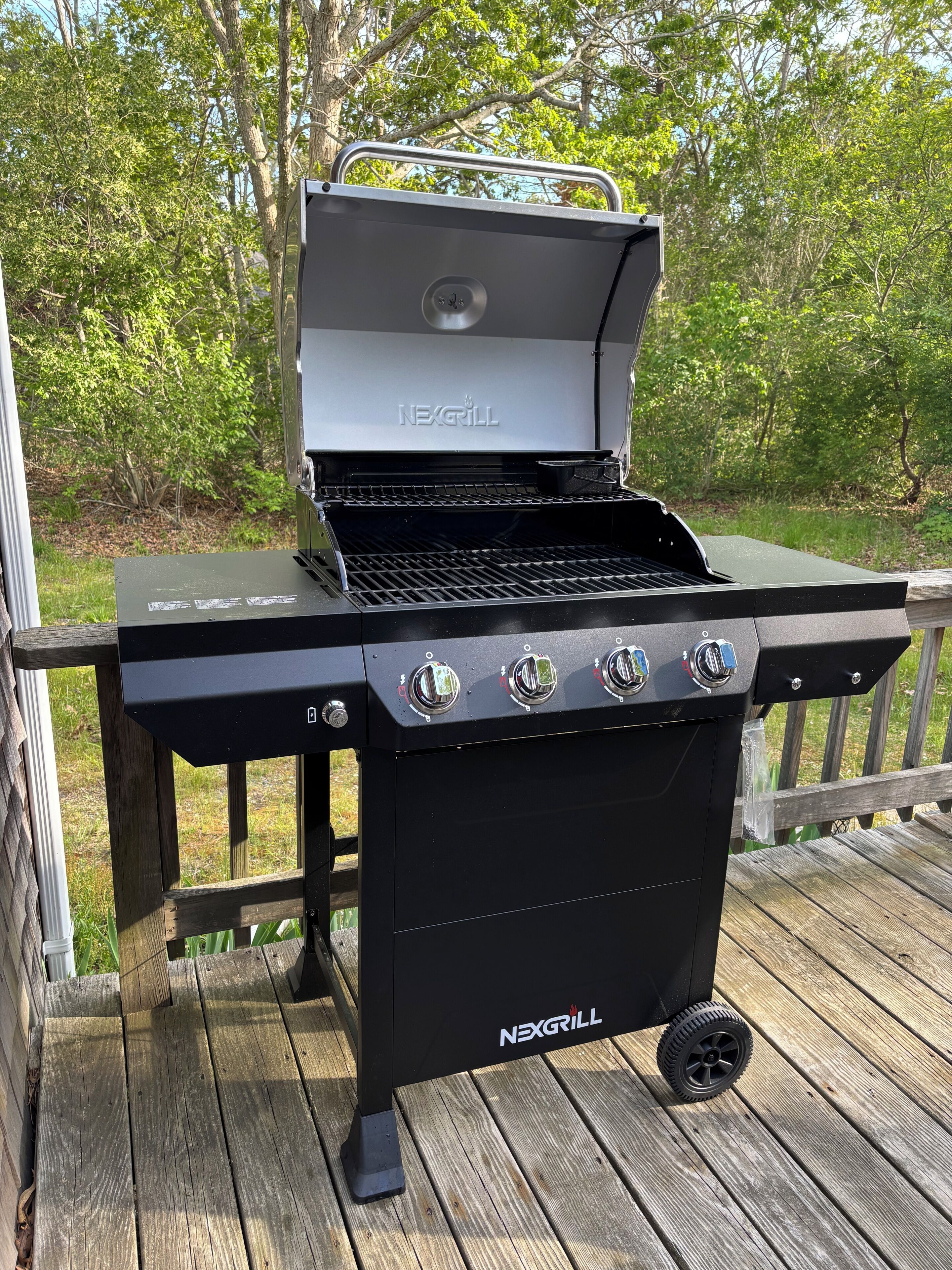 Uma churrasqueira NEXGRILL preta com quatro botões de controle e um queimador lateral, colocada sobre um deck de madeira com guarda-corpo.