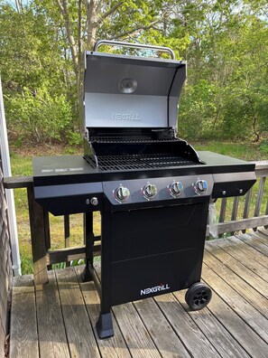 Een zwarte NEXGRILL barbecue met vier bedieningsknoppen en een zijbrander, staand op een houten terras met een reling.
