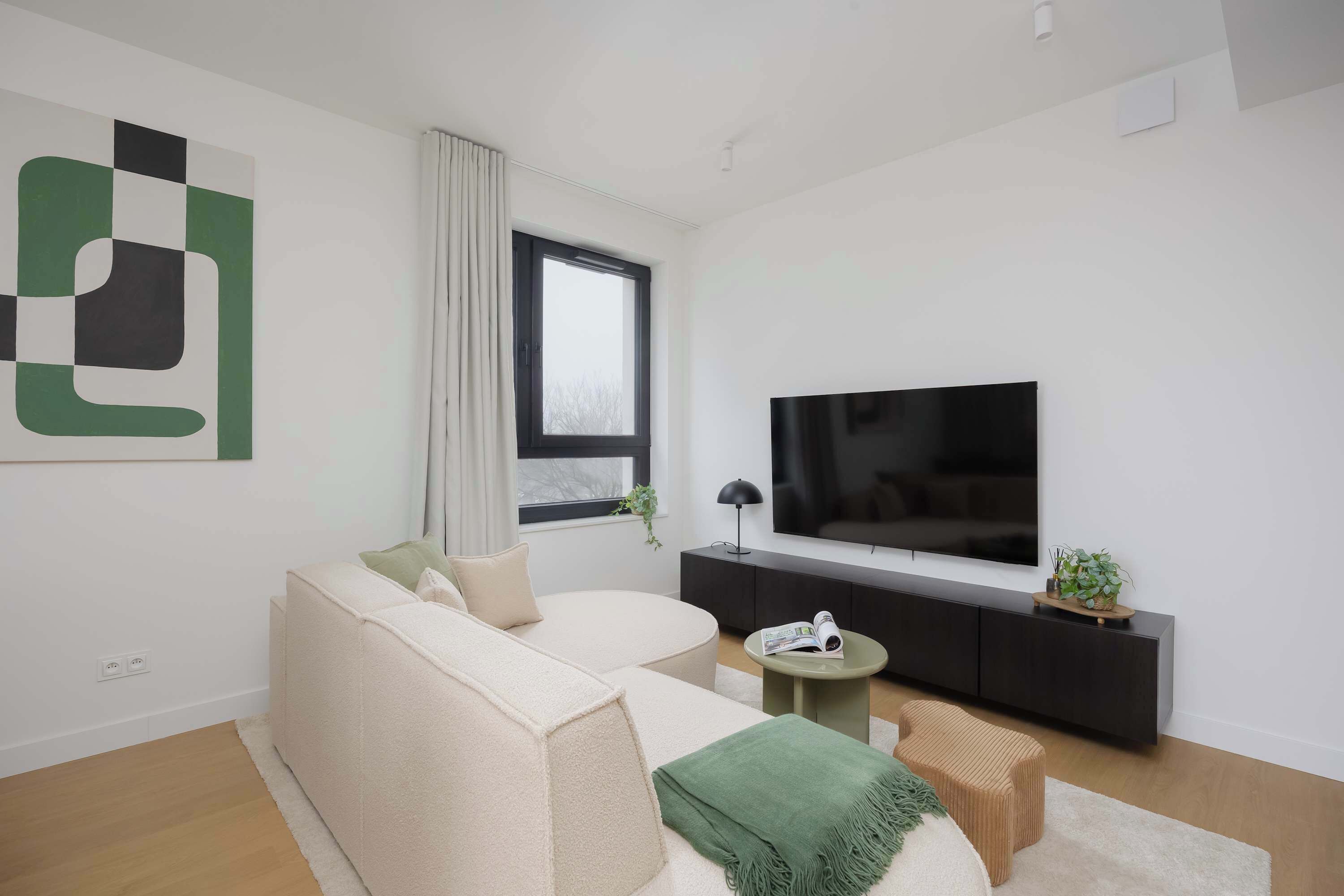 A modern living room with a large flat-screen TV, a sofa, a small round table, and a window with curtains.