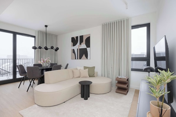 Una sala de estar moderna con un sofá modular, una mesa de comedor con sillas, una mesita auxiliar negra, una planta en maceta y un amplio ventanal con cortinas.