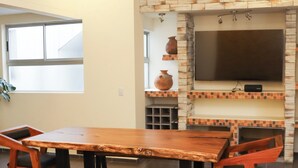 A dining area with a wooden table and chairs, a flat-screen TV mounted on a stone wall, and a window providing natural light. - An Experience of Luxury and Comfort (Medellín)