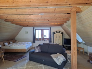 Ein gemütliches Schlafzimmer im Dachgeschoss mit Bett, Sofa, Fernseher und einem Fenster mit Vorhängen.
