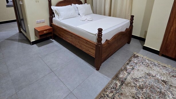 A neatly arranged bedroom with a wooden bed, a bedside table, a nightstand, a patterned rug, and a door leading to another room. - Al-mendhiry Apartment (Zanzibar)