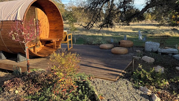 A wooden barrel-shaped structure with a deck and seating area, surrounded by a landscaped garden with rocks and plants. - Private Spa & Yurt with Sauna, Soaking Tub - Glamp & Walk to Main St (Murphys)