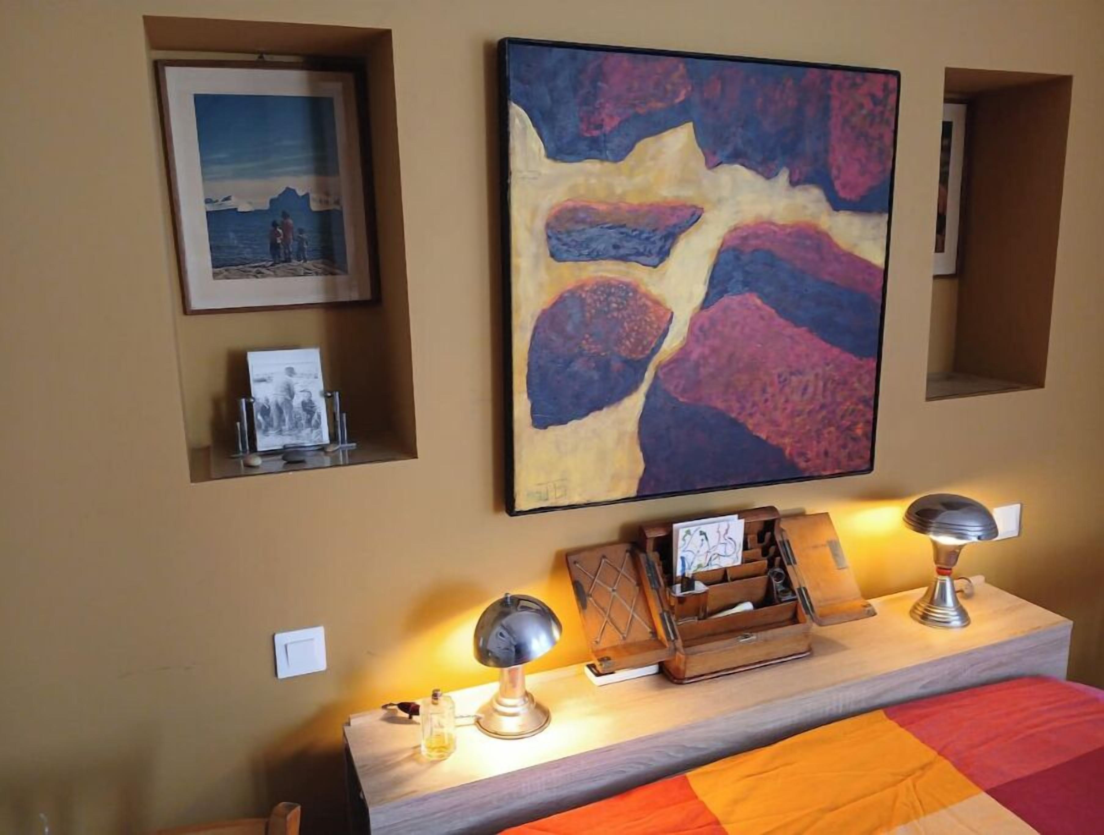 Habitación de hotel con cama, mesita de noche con lámpara, un cuadro enmarco y un gran cuadro abstracto en la pared.