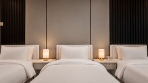 Habitación de hotel con tres camas, cada una con una mesita de noche y una lámpara. La habitación tiene un diseño moderno y una paleta de colores neutros.